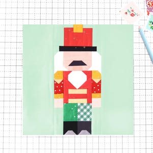 Op de afbeelding: Een kleurrijk patchwork-ontwerp van een Notenkraker-soldaat. De figuur heeft een rode jas, een zwarte hoed en wit haar. De achtergrond is lichtgroen, met een rasterpatroon. Een blauw potlood en bloemenstickers bevinden zich op de achtergrond.