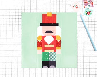 May include: A colorful, geometric patchwork design of a Nutcracker soldier. The figure has a red coat, black hat, and white hair. The background is a light green color, with a grid pattern. A blue pencil and floral stickers are in the background.