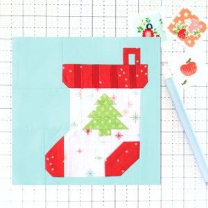 May include: A red and white Christmas stocking with a green Christmas tree on a light blue background. The stocking has a white background with a red trim and a white star pattern. The Christmas tree is green with a white outline and a white star on top.