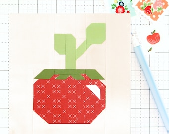 Op de afbeelding: Een stoffen blok met een geometrisch aardbeienontwerp. De rode aardbei heeft witte kruissteekdetails, groene bladeren en een groene steel. Het blok staat op een lichte achtergrond met een rasterpatroon.