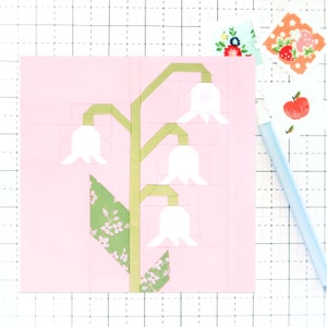 May include: A pink fabric square with a green and white appliqué of a lily of the valley flower. The flower has five white bells and green leaves.