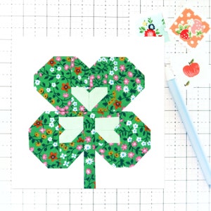 以下が含まれることがあります： 白い花の模様が入った緑色の四つ葉のクローバーが、白い背景の上に置かれています。