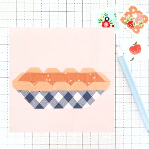 May include: A patchwork quilt block featuring a pie design. The pie crust is made of blue and white gingham fabric. The pie filling is made of orange fabric with white polka dots. The block is on a pink background.