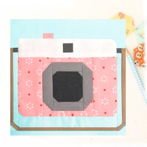 May include: A pink and white camera with a gray lens and a black viewfinder. The camera is surrounded by a light blue border and a brown border.