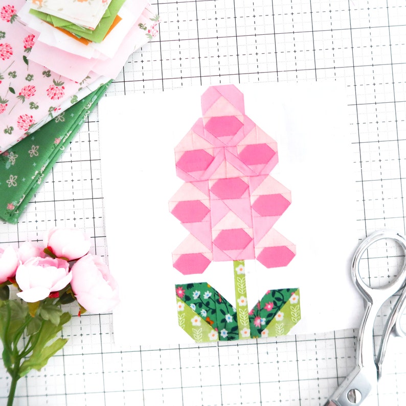 May include: A pink and green fabric flower block, made with patchwork squares, on a white grid background.
