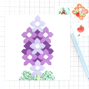 May include: A handmade quilt block featuring a stylized purple flower. The flower is composed of various shades of purple fabric, with white accents. Green and white floral fabric forms the base. The block is on a white background.