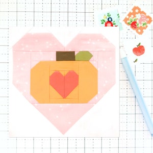 Può includere: Un blocco di tessuto a forma di cuore rosa con un blocco di zucca arancione più piccolo all'interno. La zucca ha un piccolo cuore rosa al centro e uno stelo marrone con una foglia verde.