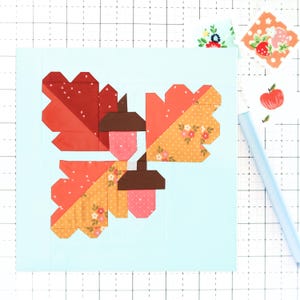 Könnte beinhalten: Ein farbenfroher Patchwork-Block mit drei stilisierten Eicheln und Blättern. Die Blätter sind in Orange- und Rottönen gehalten, einige Abschnitte sind mit kleinen floralen Mustern versehen. Die Eicheln haben braune Kappen und rosa Körper, vor einem hellblauen Hintergrund.