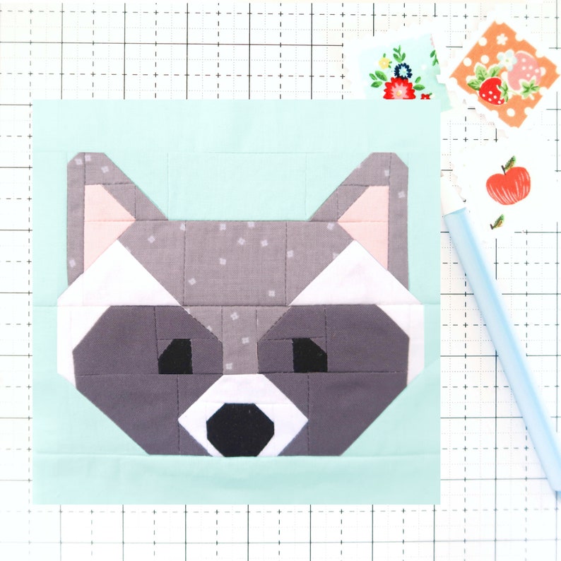 May include: A patchwork quilt block featuring a raccoon face in shades of gray, pink, white, and black. The block is set against a light blue background.