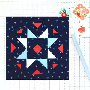 May include: A square fabric block with a starburst design. The star is composed of red, white, and light blue triangles and a red diamond on a navy blue floral background. Small floral stickers and a light blue pen are also visible.