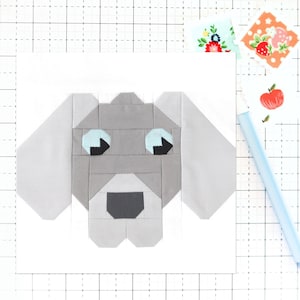 May include: A geometric fabric dog face in shades of gray, light blue, and black, with large ears and a friendly expression. The dog is centered on a white background, with colorful floral and fruit-themed fabric scraps and a light blue pen nearby.