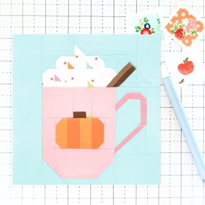 May include: A pink mug with a brown stick and white whipped cream on top. A small orange pumpkin is inside the mug. The mug is on a light blue background.
