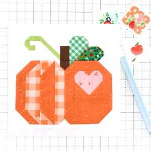 May include: A fabric pumpkin block with an orange and white gingham pattern on the left side and a heart shape on the right. The pumpkin has a green stem and a brown stem base. The background includes fabric scraps and a light blue pen.
