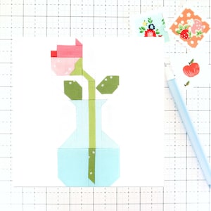 May include: A pink and green fabric flower in a light blue vase. The flower is made of patchwork fabric squares and the vase is made of patchwork fabric rectangles.