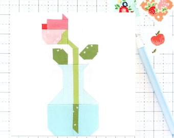May include: A pink and green fabric flower in a light blue vase. The flower is made of patchwork fabric squares and the vase is made of patchwork fabric rectangles.