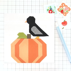 Op de afbeelding: Een zwarte vogel die op een oranje pompoen met groene bladeren zit. De vogel heeft een grijze vleugel en een gele snavel. De pompoen is gemaakt van stof en heeft een patchwork-ontwerp.