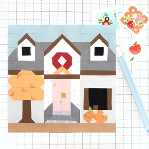 May include: A patchwork quilt block depicting a house with a pink door, a brown tree with orange leaves, and a wreath on the front door. The house has a gray roof and brown trim. The quilt block is made of various fabrics in shades of gray, brown, pink, orange, and white.