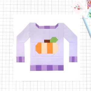 May include: A light purple sweater with a pumpkin design. The pumpkin is orange and cream with a green leaf and brown stem. The sweater is made of fabric and is on a white surface.