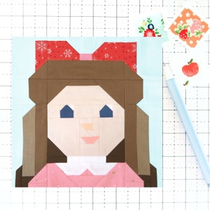 Puede incluir: Un bloque de colcha de patchwork que representa a una niña con cabello castaño, un lazo rojo y un vestido rosa. El bloque está hecho de varias telas con diferentes patrones y colores.