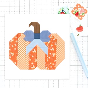 May include: A geometric patchwork pumpkin design. The pumpkin is composed of orange fabric with white floral and gingham patterns. It features a blue bow, a brown stem, and is set against a white background. Additional fabric swatches and a blue pen are visible.