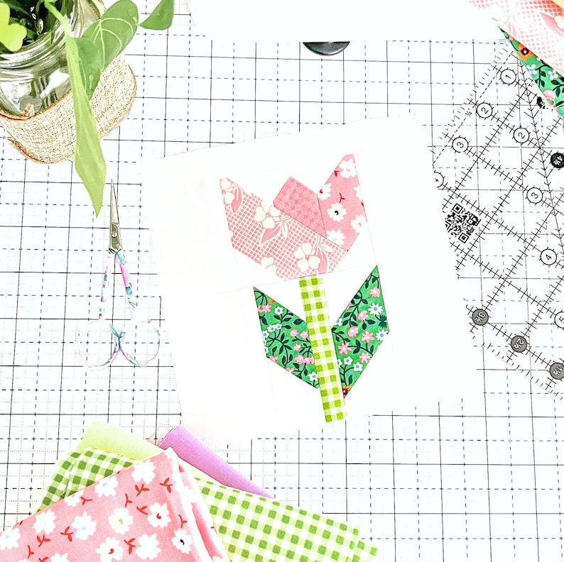 May include: A patchwork tulip flower block made from pink and green fabric with a white background. The fabric has a floral pattern. The block is on a white cutting mat with a grid pattern.
