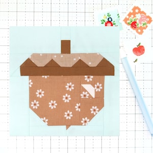 Op de afbeelding: Een bruin en wit stoffen blok in de vorm van een eikel met een bruine steel. Het blok bevindt zich op een lichtblauwe achtergrond.