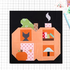 Peut inclure: Un bloc de courtepointe en tissu en forme de citrouille sur fond noir. La citrouille présente des sections en tissu à motifs orange, rose et vert, incluant une silhouette de chat noir, une étagère, une petite citrouille et une lampe.