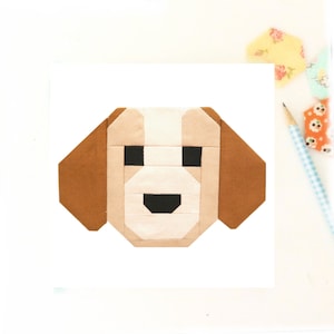 May include: A geometric dog face design in shades of tan, brown, and white, with black eyes and a mouth. The artwork is likely a quilt block or fabric design, with a clean, modern aesthetic. The dog's ears are a darker brown.