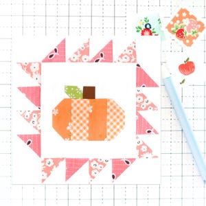 Könnte beinhalten: Ein Patchwork-Kürbis mit einem grünen Stiel und orangefarbenen, weißen und rosa Stoffquadraten ist von einem rosa-weißen Blumenrand umgeben. Der Rand besteht aus kleinen Dreiecken.