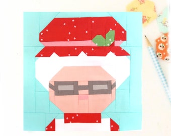 Op de afbeelding: Een patchwork quiltblok met een cartoon oma met een rode hoed, wit haar en een bril. Het blok is gemaakt van verschillende stoffen in tinten rood, wit, roze, groen en blauw.
