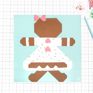 May include: A paper pieced gingerbread person quilt block. The block features a brown gingerbread person with a pink bow, a white dress with colorful sprinkles, and pink accents. The background is light blue.
