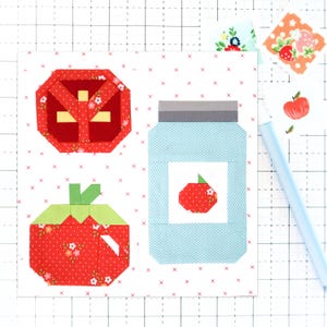 Puede incluir: Una obra de arte textil con diseños geométricos de un tomate, una fresa y un tarro de mermelada. El tomate y la fresa son rojos con estampados florales, mientras que el tarro es azul claro con una etiqueta blanca. Un bolígrafo azul claro está a la derecha.
