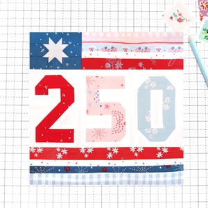 May include: A patchwork quilt block with a blue and white star, red stripes, and the number "250". The design includes floral and snowflake patterns. A light blue pencil and small fabric squares are in the background.