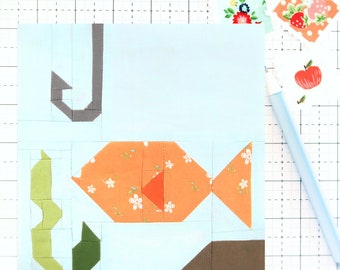 Könnte beinhalten: Ein hellblaues Patchwork-Quadrat mit einem orangefarbenen Fisch mit weißen Blumenakzenten. Der Fisch ist von grünen und braunen geometrischen Formen umgeben. Ein hellblauer Stift und dekorative Aufkleber mit Blumen- und Fruchtdesigns sind ebenfalls sichtbar.