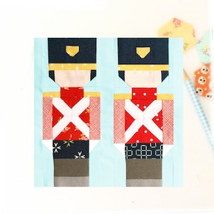 May include: Two toy soldier quilt blocks, each with a red, white and blue patterned fabric. The blocks are made of different fabrics, including a red fabric with white polka dots, a red fabric with white and green stars, and a navy blue fabric with white stars.