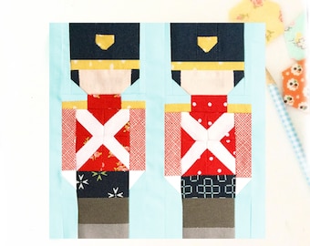 May include: Two toy soldier quilt blocks, each with a red, white, and blue patterned fabric. The blocks are made of different fabrics, including a red fabric with white polka dots, a red fabric with white and green stars, and a navy blue fabric with white stars.