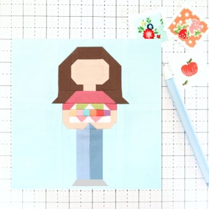 May include: A blue background with a brown-haired person wearing a pink shirt and blue jeans. The person is holding a colorful quilt in their hands.