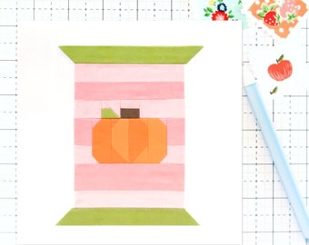 May include: A pink and green fabric applique of a pumpkin with a brown stem. The pumpkin is on a pink and white striped background.