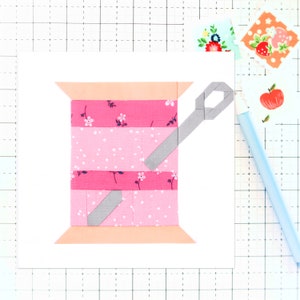 May include: A pink and white fabric spool with a gray needle threaded through it. The spool is on a white background.
