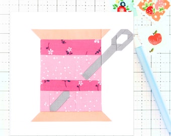 May include: A pink and white fabric spool with a gray needle threaded through it. The spool is on a white background.