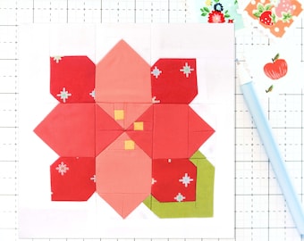 Peut inclure: Un bloc de fleur en tissu rouge, rose et vert sur un fond blanc. Le bloc est composé de plusieurs petits carrés et triangles, et il est cousu avec un fil blanc.