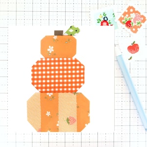 May include: A three-tiered patchwork pumpkin made from orange fabric with different patterns. The top tier has a floral pattern, the middle tier has a gingham pattern, and the bottom tier has a floral and stripe pattern. The pumpkin has a brown stem and a green leaf.