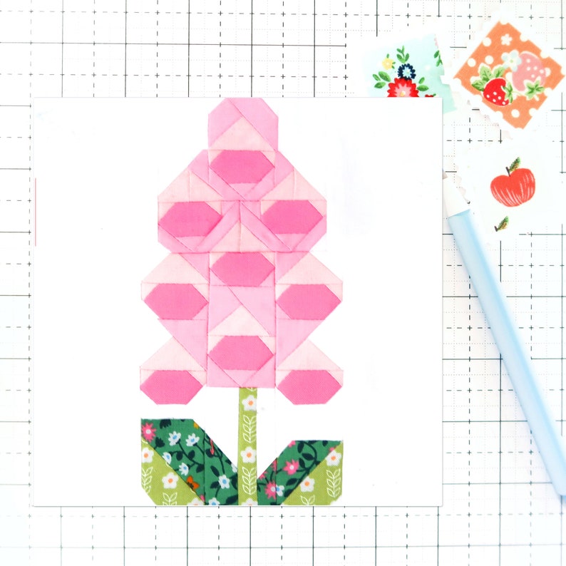 May include: A pink and green fabric flower applique with a stem and leaves. The flower is made of multiple pink fabric squares sewn together to create a layered effect.