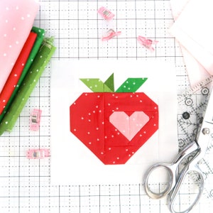 Peut inclure: Une fraise en tissu rouge et vert avec un cœur rose au centre, sur un fond blanc avec un motif quadrillé.