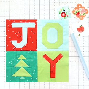 Op de afbeelding: Een kleurrijk patchwork quilt vierkant met het woord "JOY" in vette letters. De letters zijn rood, groen en lichtblauw, met een kerstboomontwerp. Kleine bloemenstof vierkanten en een lichtblauwe pen zijn ook zichtbaar.