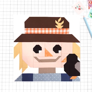 May include: A patchwork scarecrow design with a brown hat, orange gingham hatband, and a black bird. The scarecrow has a light skin tone, blonde hair, and a blue and white polka dot shirt. A light blue pencil and floral stickers are in the background.