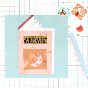 May include: A light blue fabric square with a pink book sewn on top. The book has a title that reads "Tales of the Wild West" and a moon and floral pattern on the cover.