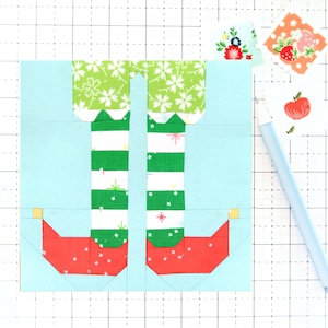 May include: A light blue background with a red, green, and white elf leg applique. The applique is made of fabric and has a scalloped edge. The applique is on a gridded surface.