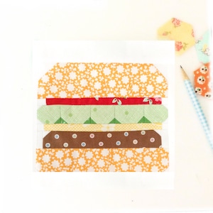 Könnte beinhalten: Ein farbenfrohes Patchwork-Design aus Stoff, das einen Hamburger mit einem gelben Brötchen, einer roten Tomate, grünem Salat, einem braunen Patty und einem gelben unteren Brötchen darstellt.