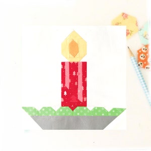 May include: A red candle with a white and pink Christmas tree pattern, a yellow flame, and a green and white wreath base.